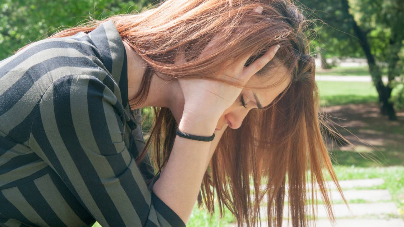 Depressed girl sitting on bench in park. Side view of frustrated young woman with closed eyes covering ears with hands and holding head. Depressive syndrome concept