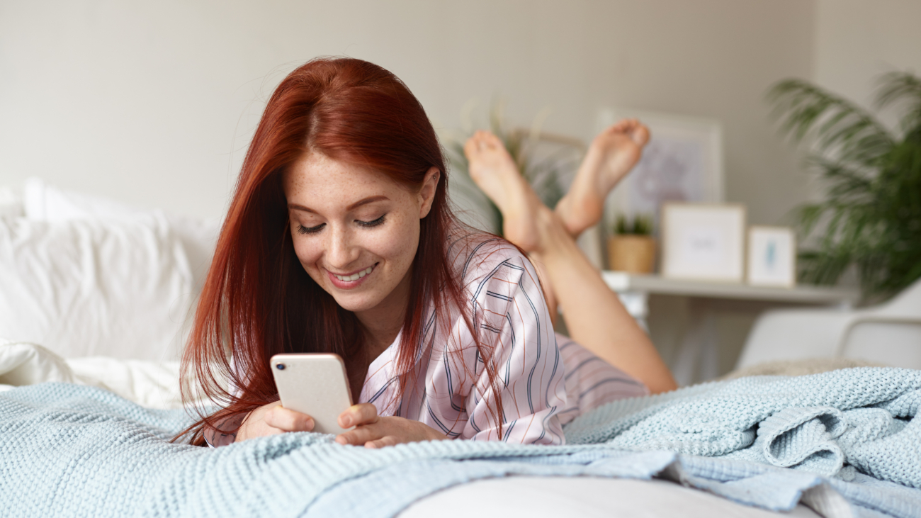 Cheerful playful young redhead female in pajamas lying on bed in her room with feet in the air, smiling joyfully while flirting with her boyfriend, sending him text message online using mobile phone